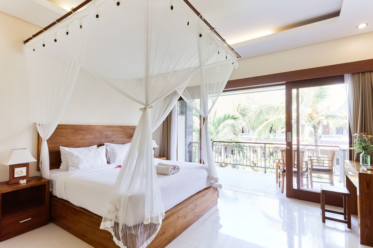 A bedroom with a four-poster bed, a nightstand, a desk with a chair, and a balcony with a view of palm trees.