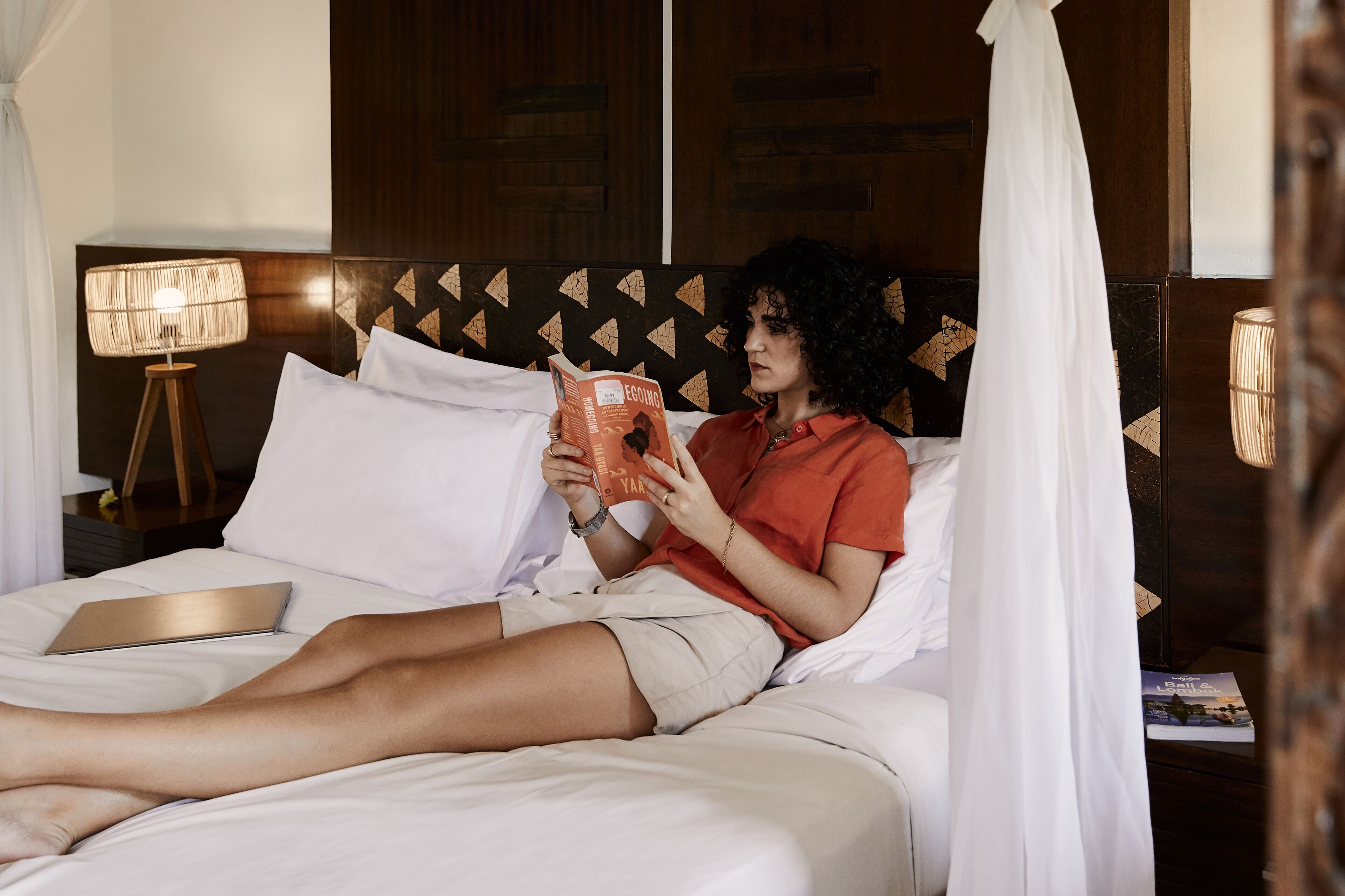 A person lying in bed reading a book, with a laptop on the bed and a lamp beside the bed.