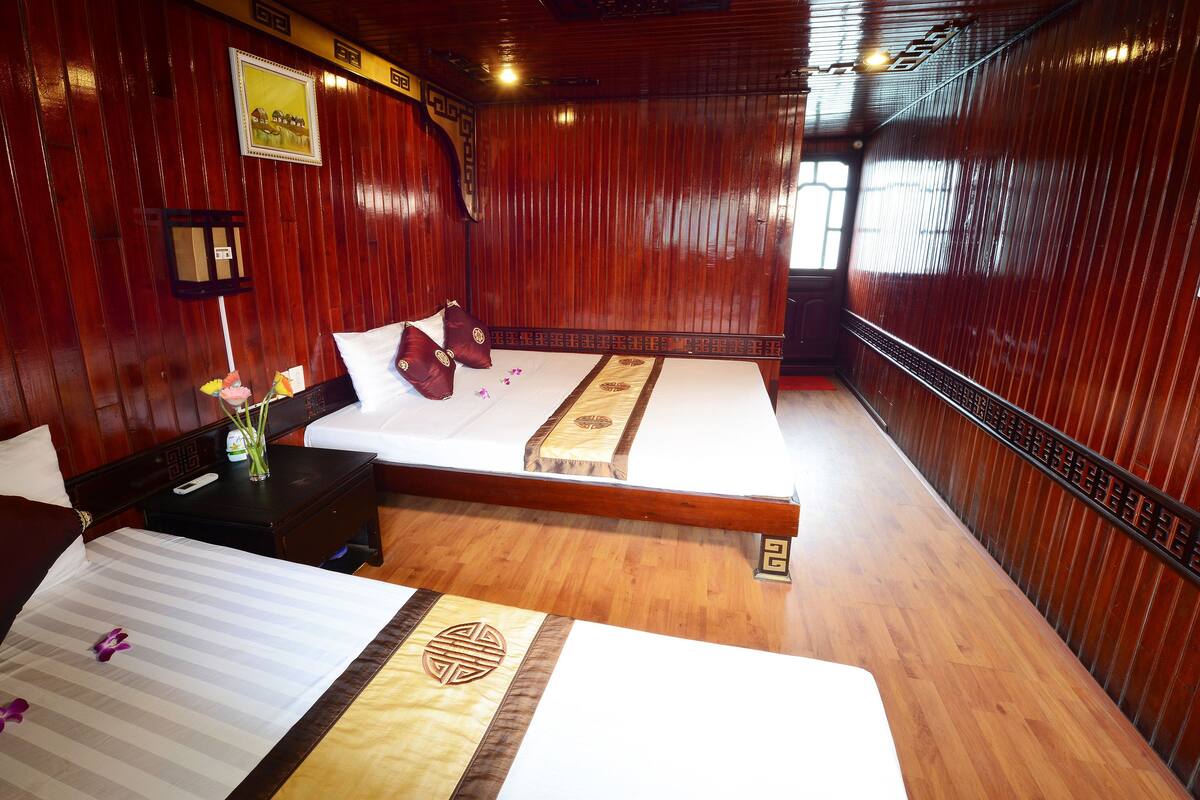 Quarto de hotel com duas camas, painéis de madeira, uma pequena mesa de cabeceira com um vaso e controle remoto, e um travesseiro decorativo.