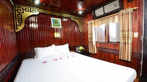 A neatly made bed with white linens and pink flower petals, a wooden headboard with decorative carvings, and a window with curtains. - Imperial Legend Cruise Halong (Tuan Chau)