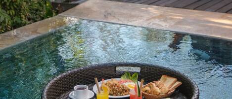 Una mesa de comedor flotante con alimentos para el desayuno junto a una piscina, rodeada de exuberante vegetación y una terraza de madera.