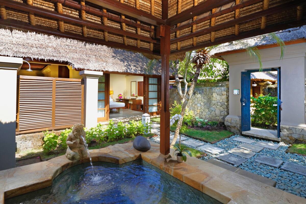 A traditional thatched-roof structure with a stone pathway leading to a pool area, surrounded by lush greenery and a stone wall.