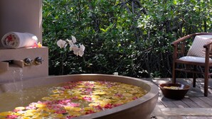 Uma banheira cheia de água e pétalas de flores, uma toalha e uma cadeira em um deck de madeira, com vegetação exuberante ao fundo.