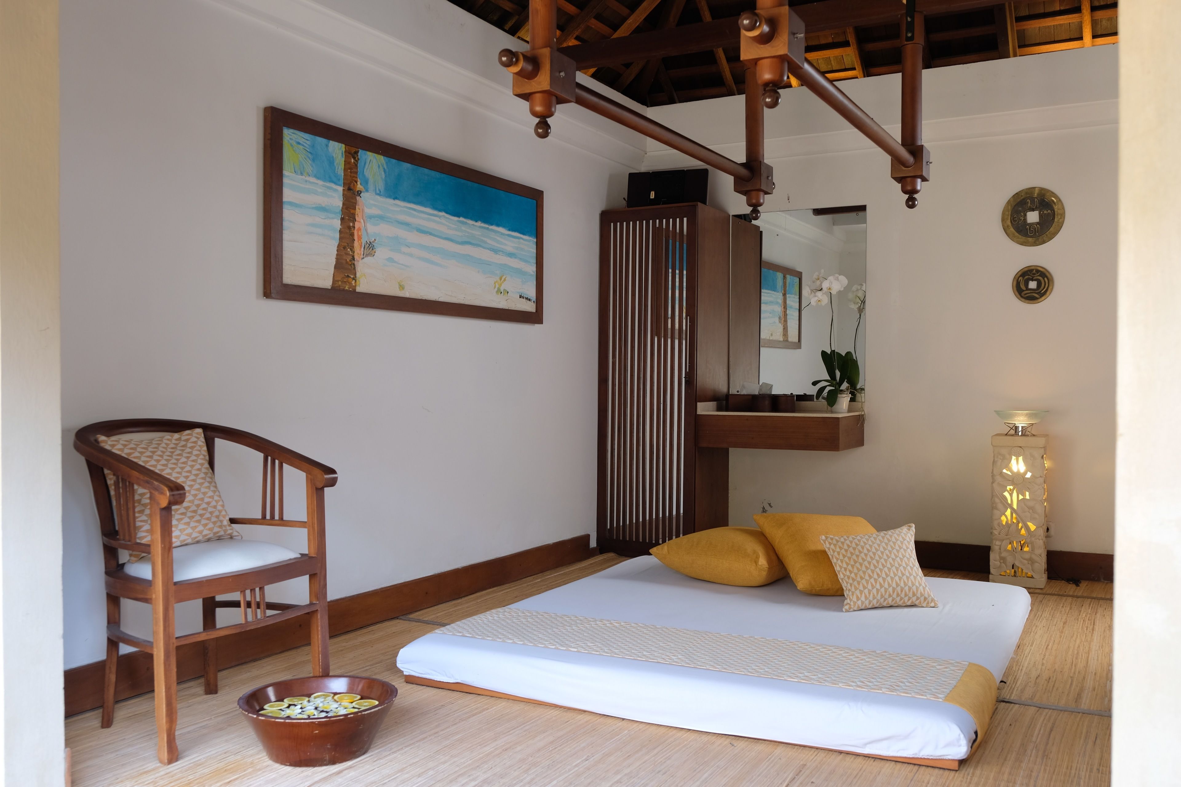 A bedroom with a bed, a wooden chair, a bowl, a painting, a mirror, and a wall clock.