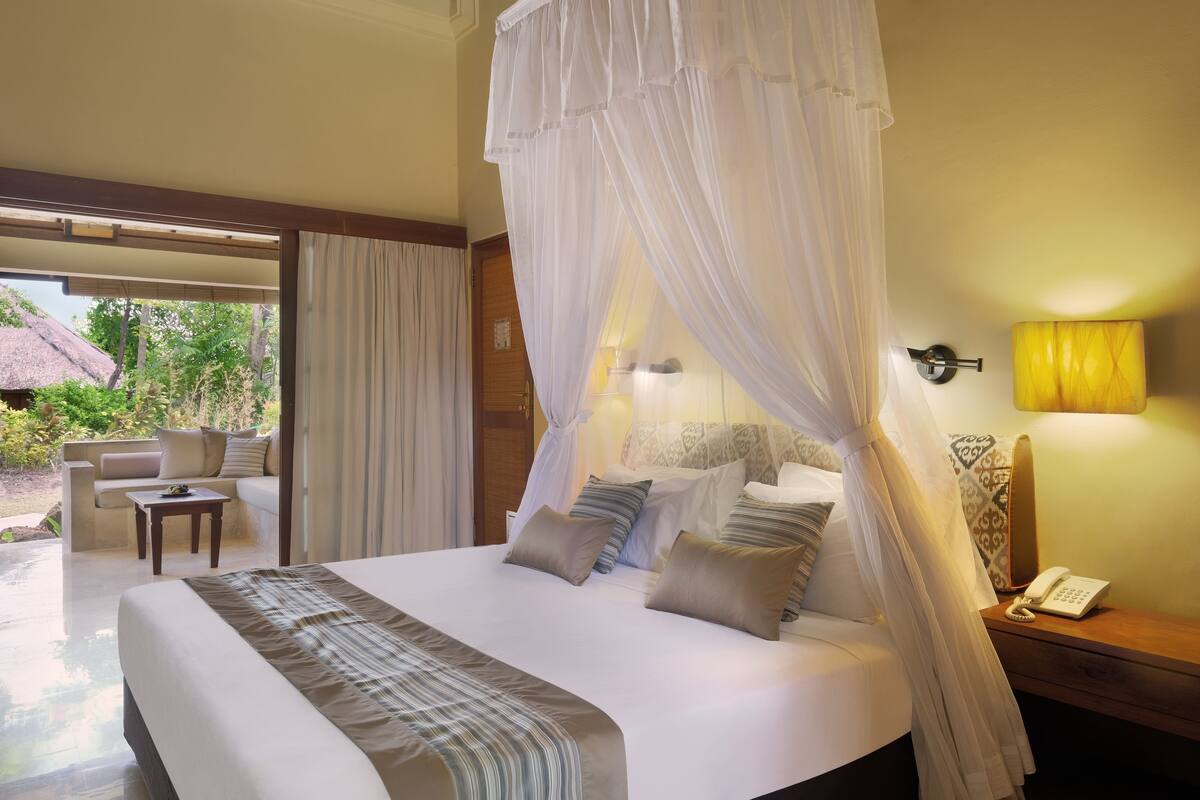 A bedroom with a bed, bedside table, and a view of an outdoor area with a sofa and a thatched roof structure.