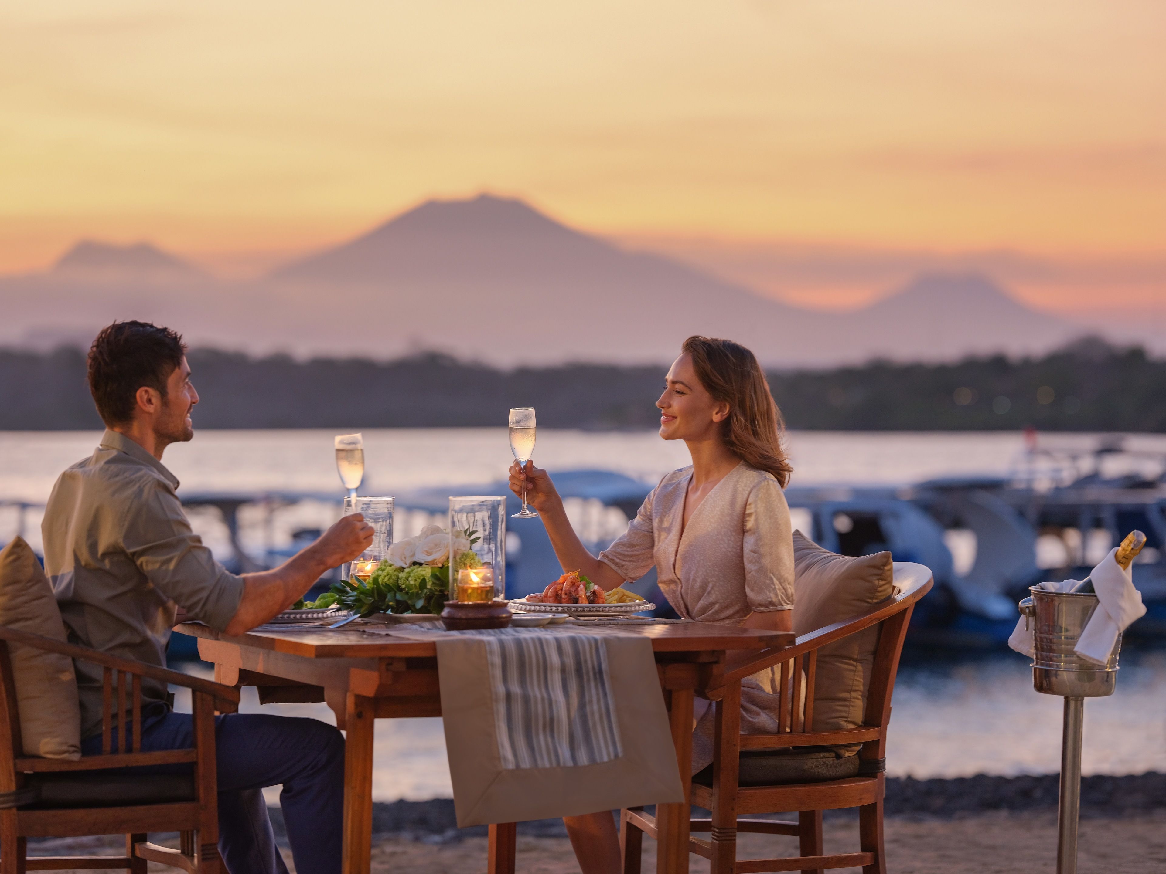 Un couple est assis à une table en bois, dînant, avec en arrière-plan une vue sur des bateaux et des montagnes.