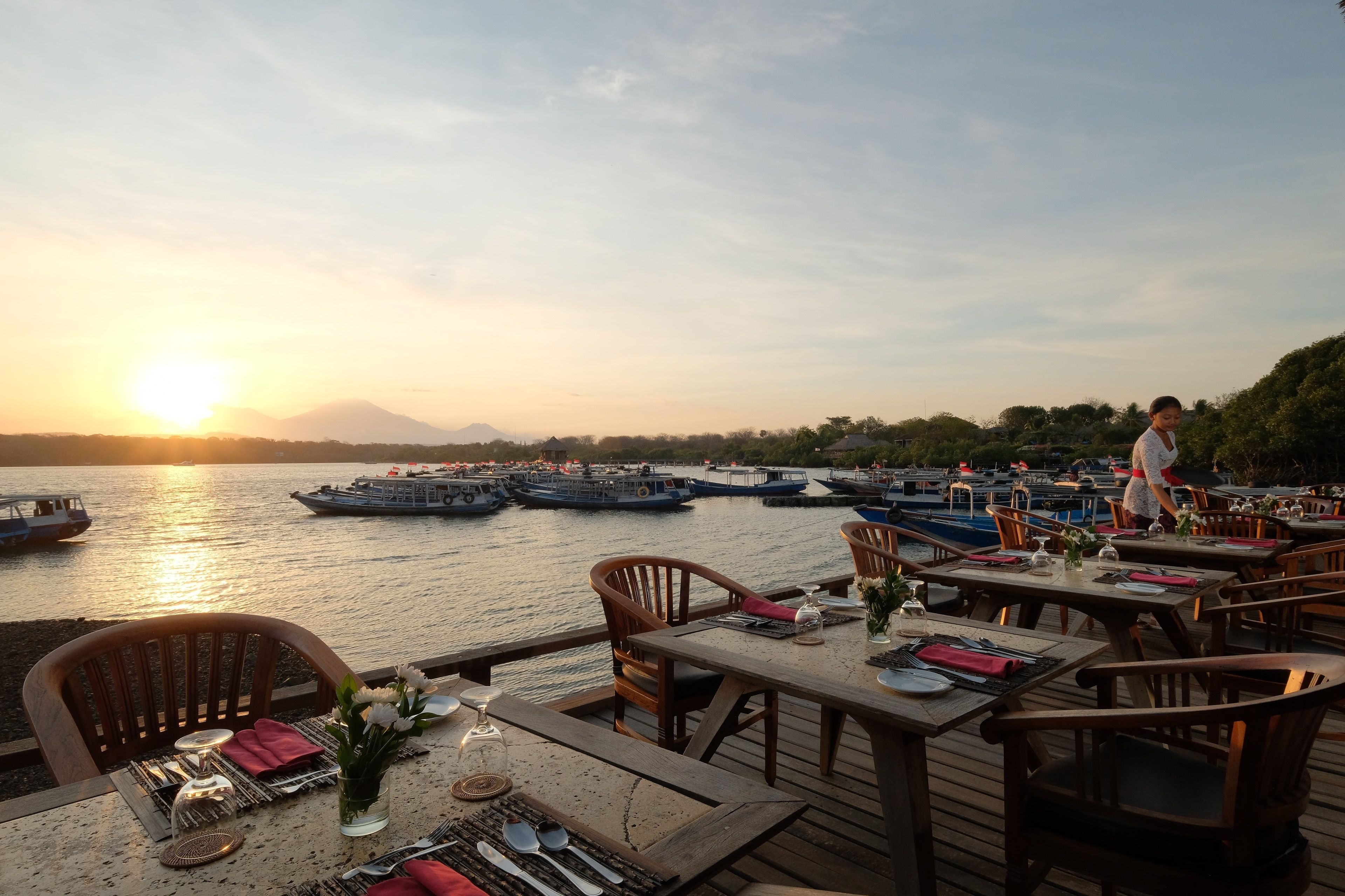Un restaurante frente al agua, con mesas preparadas para comer, con vista a una masa de agua tranquila donde se ven barcos y una montaña al fondo.