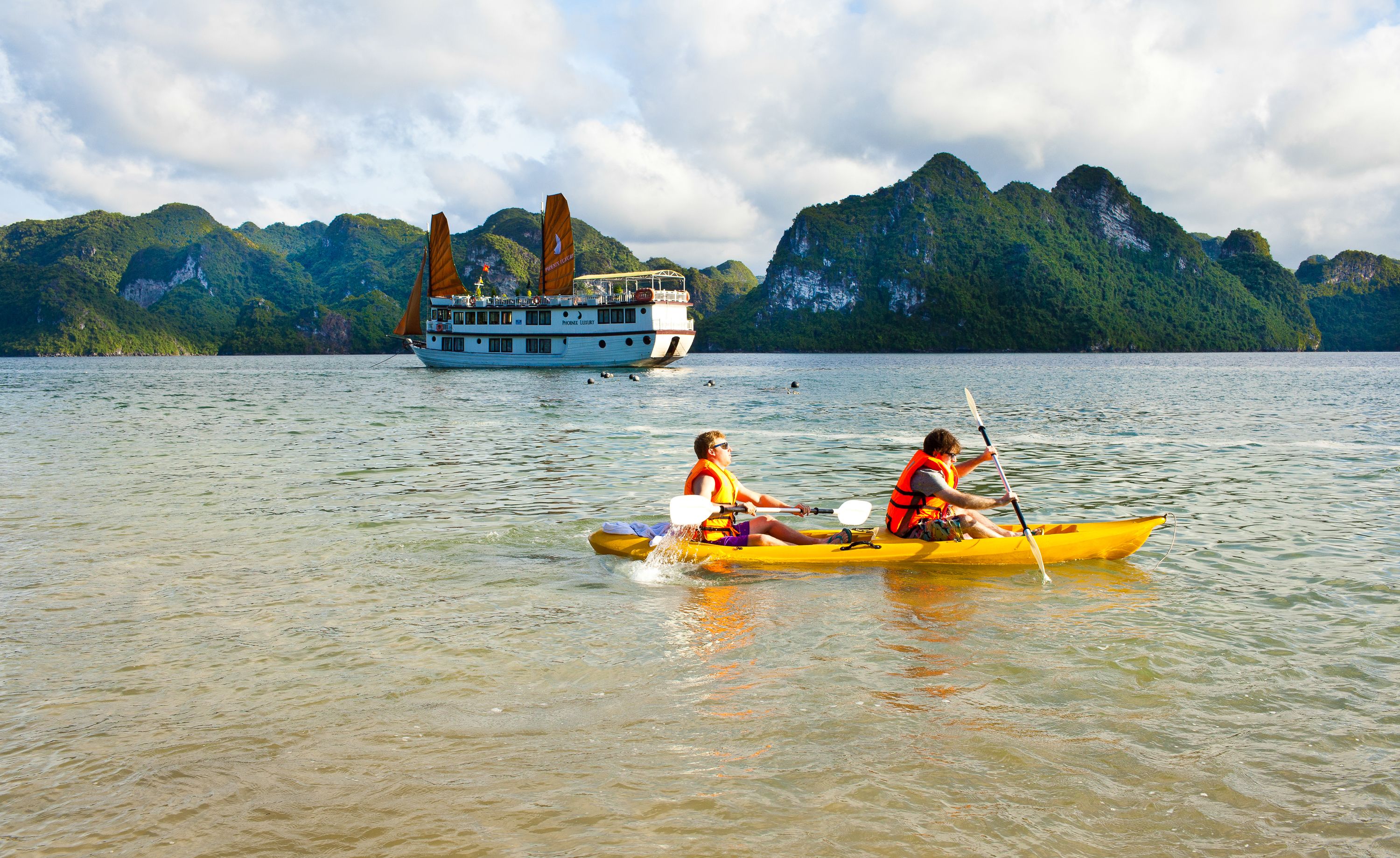 Une personne dans un kayak jaune sur un lac calme, avec un grand bateau et des montagnes en arrière-plan.