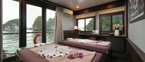 A room with two beds, large windows with a view of a body of water and greenery, a desk with a lamp, and a life preserver on the window sill.