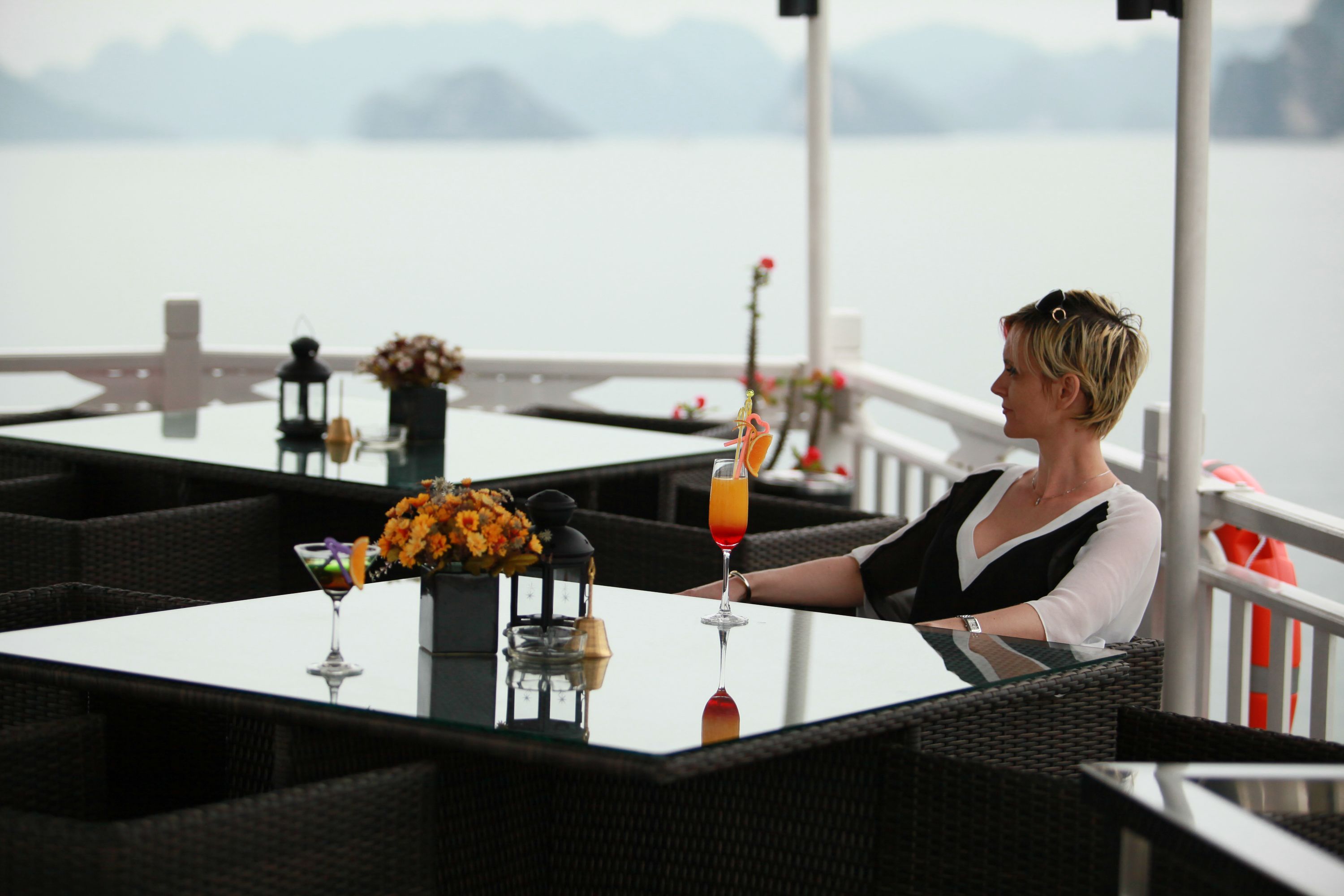 A woman seated at a table with a drink, overlooking a body of water.