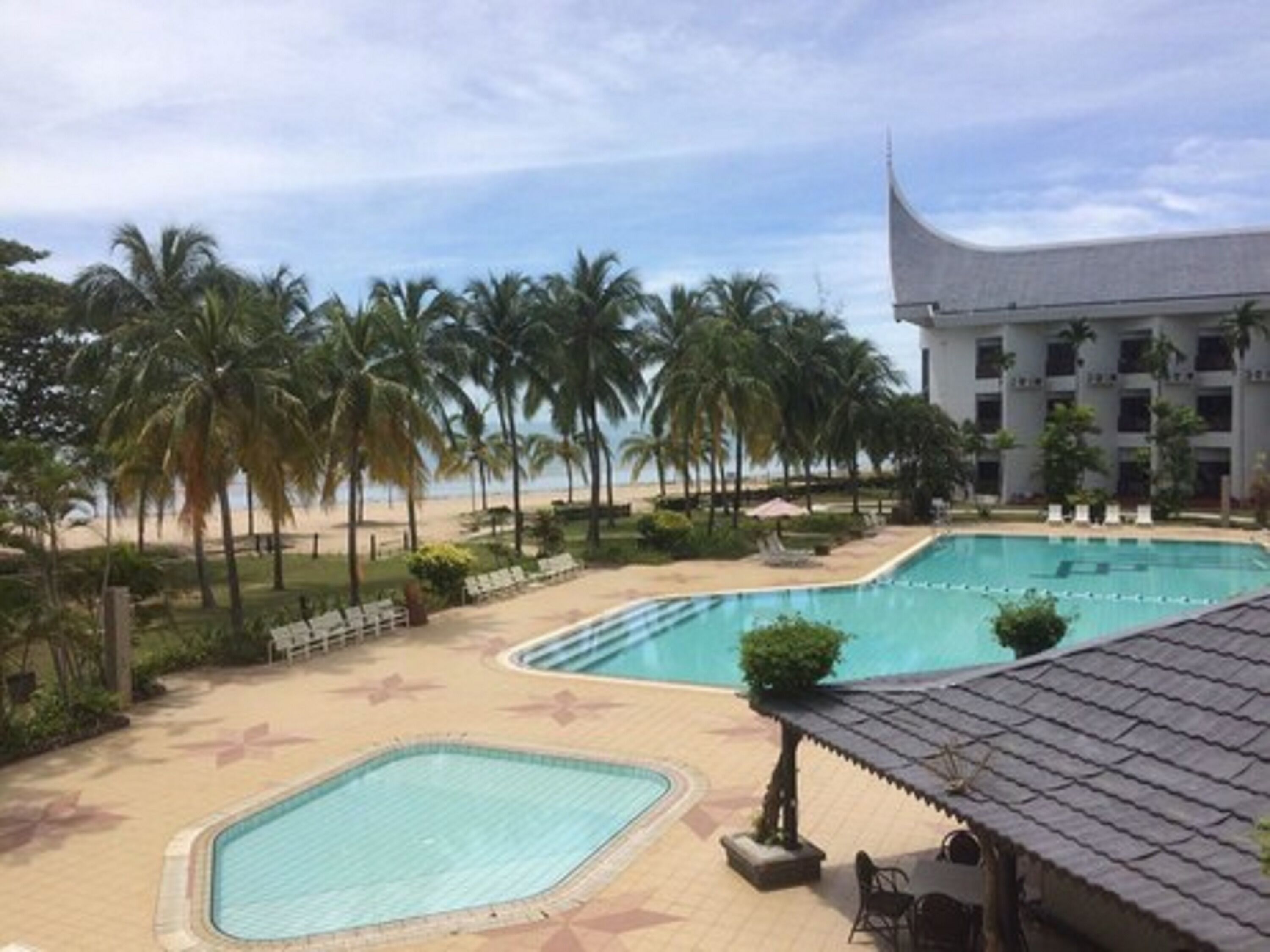 A pool area with palm trees, a beach, and a building with a unique roof design.