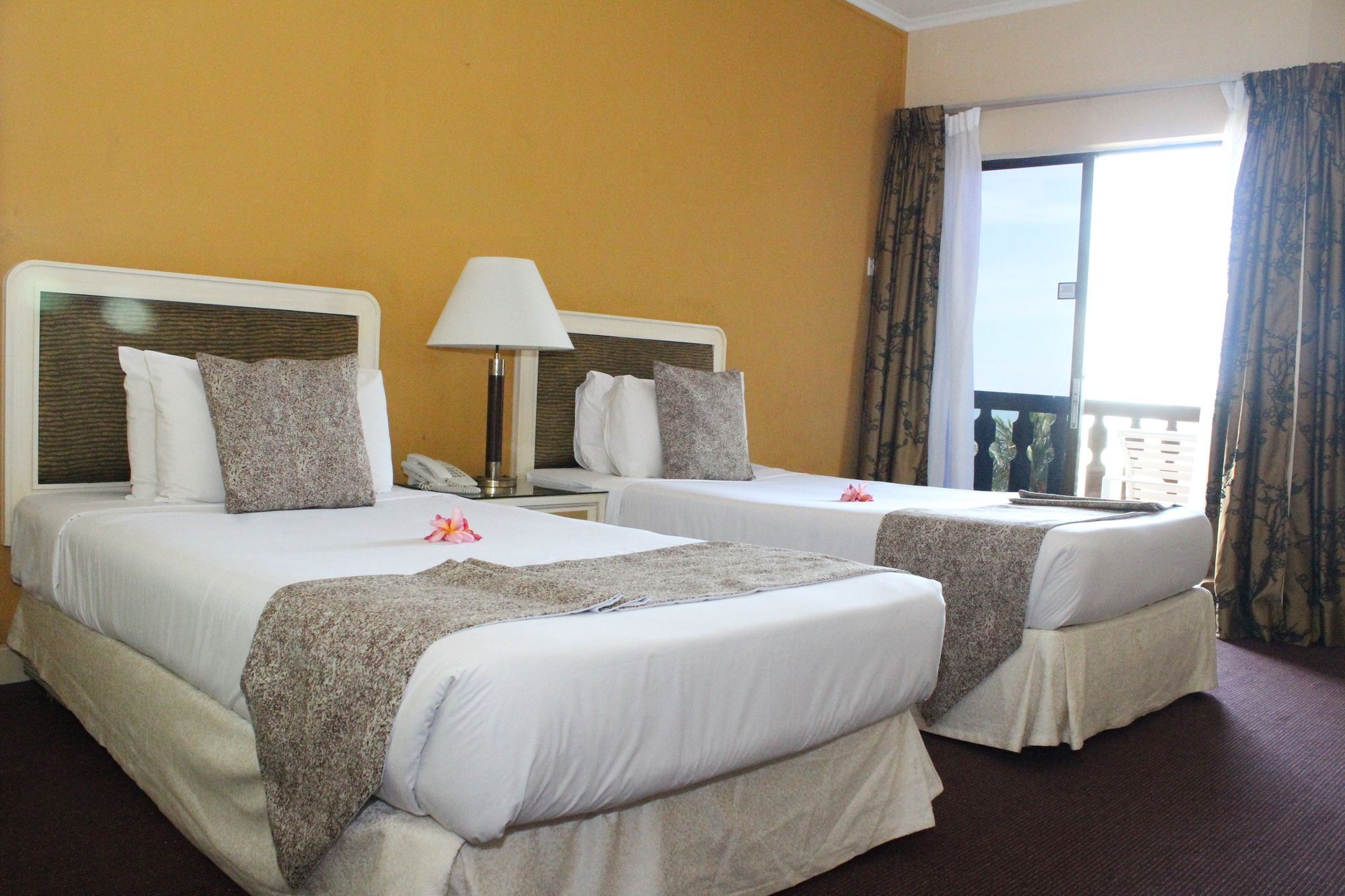 A hotel room with two beds, a nightstand with a lamp, a phone, and a balcony with curtains.