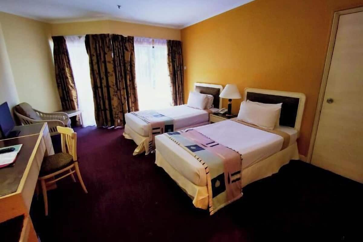 Ein Hotelzimmer mit zwei Betten, einem Schreibtisch mit Stuhl, einer Lampe und einem Fenster mit Vorhängen.
