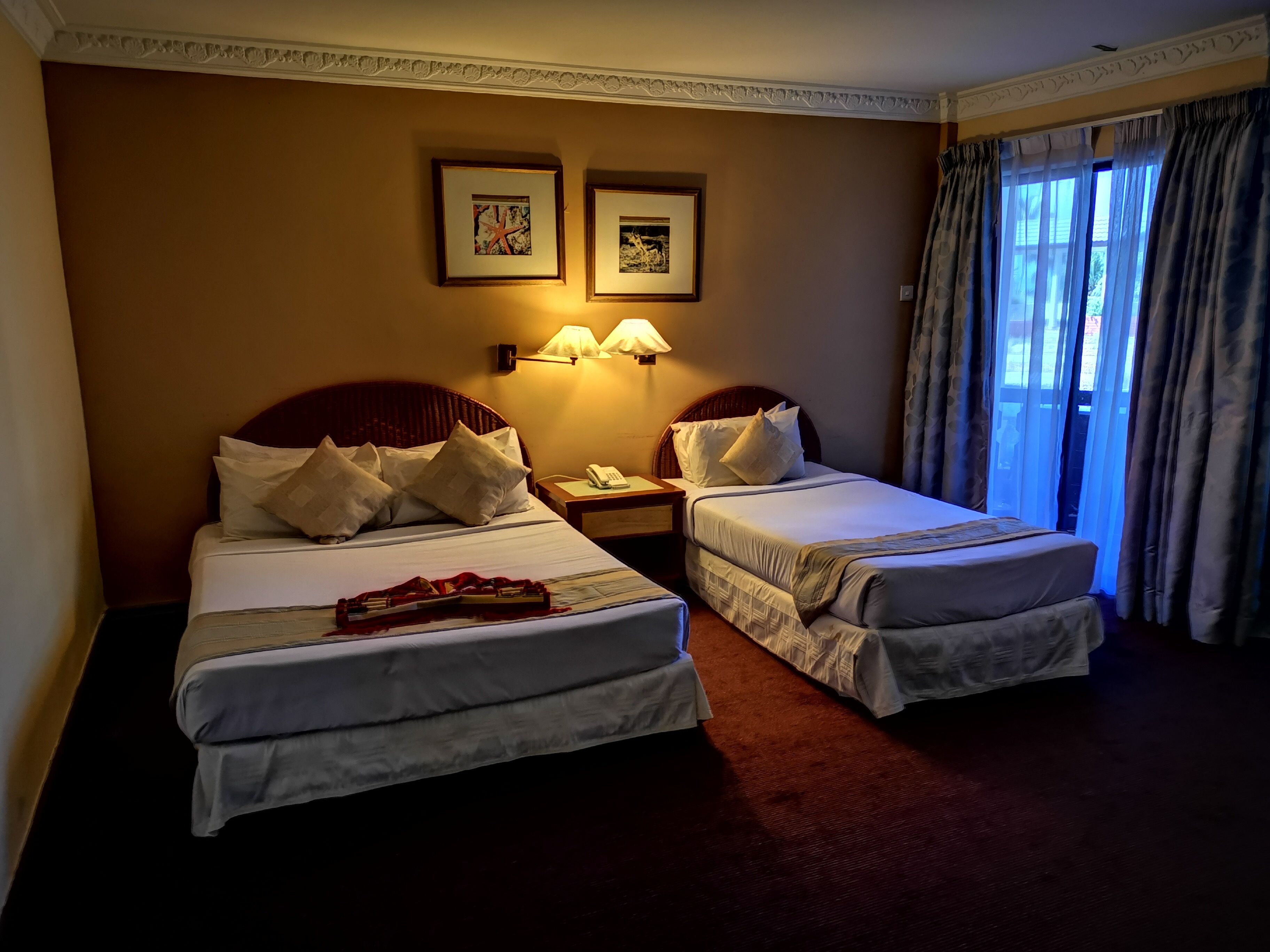Ein Hotelzimmer mit zwei Einzelbetten, einem Nachttisch mit Lampe und einem Fenster mit Vorhängen.