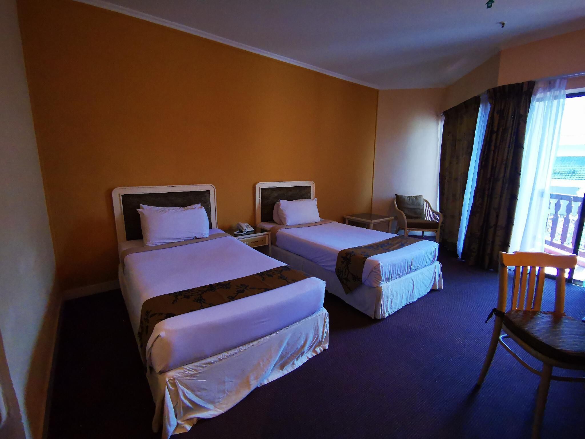 Ein Hotelzimmer mit zwei Betten, einem Sessel, einem kleinen Tisch und einem Balkon mit Vorhängen.
