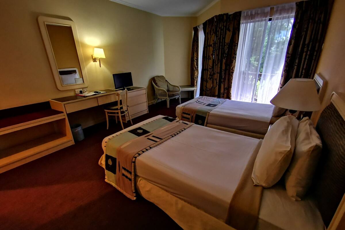 Ein Hotelzimmer mit zwei Betten, einem Schreibtisch, einem Stuhl, einem Spiegel und einem Fenster mit Vorhängen.