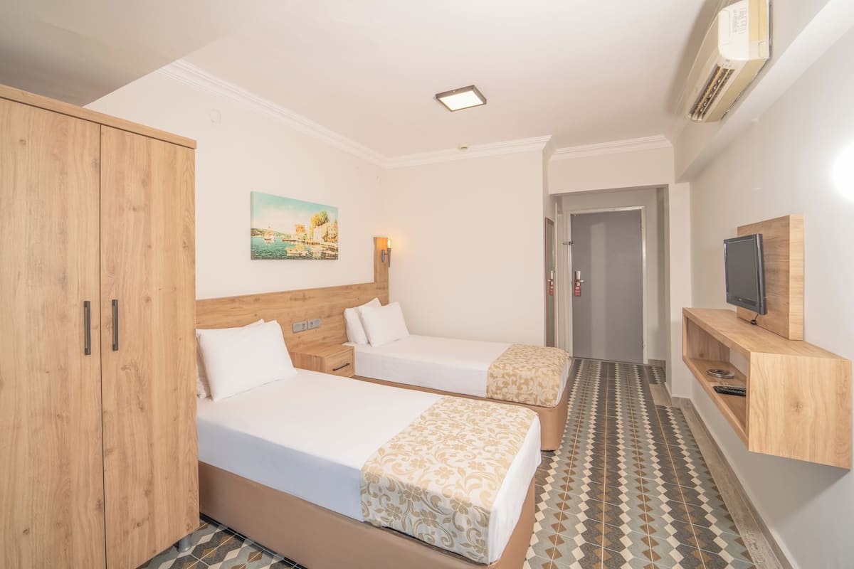 A hotel room with two beds, a wooden wardrobe, a painting on the wall, a television on a wooden shelf, and a patterned floor.