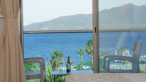 Una habitación con un ventanal grande que ofrece vistas al mar y a las montañas. Hay una mesa con una botella y un vaso, y dos sillas.