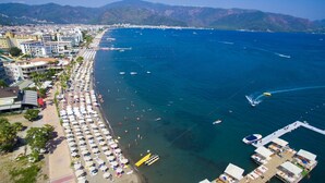 Vista aérea de una ciudad costera con una playa extensa bordeada de sombrillas y tumbonas, un puerto deportivo con barcos y edificios a lo largo de la costa.
