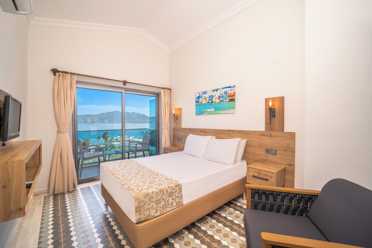 A hotel room with a large bed, a desk, a chair, a TV, a balcony with a view of the sea, and a painting on the wall.
