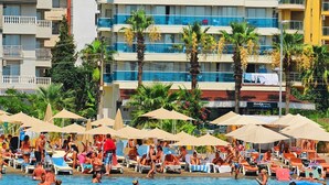 Zona de piscina de hotel con múltiples tumbonas y sombrillas, rodeada de palmeras y un edificio con balcones.