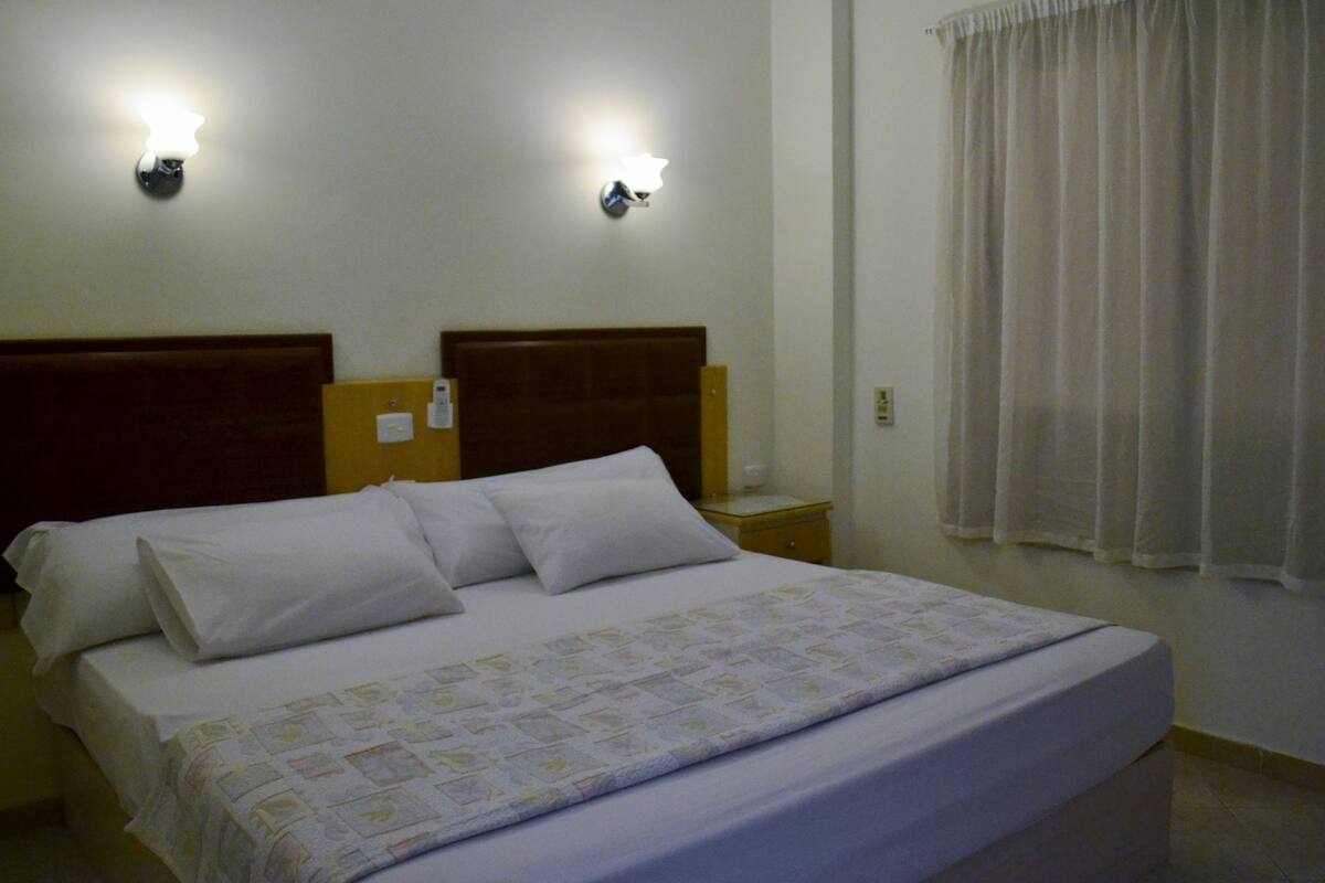 A hotel room with a large bed, two wall-mounted lights, a nightstand, and a window with curtains.
