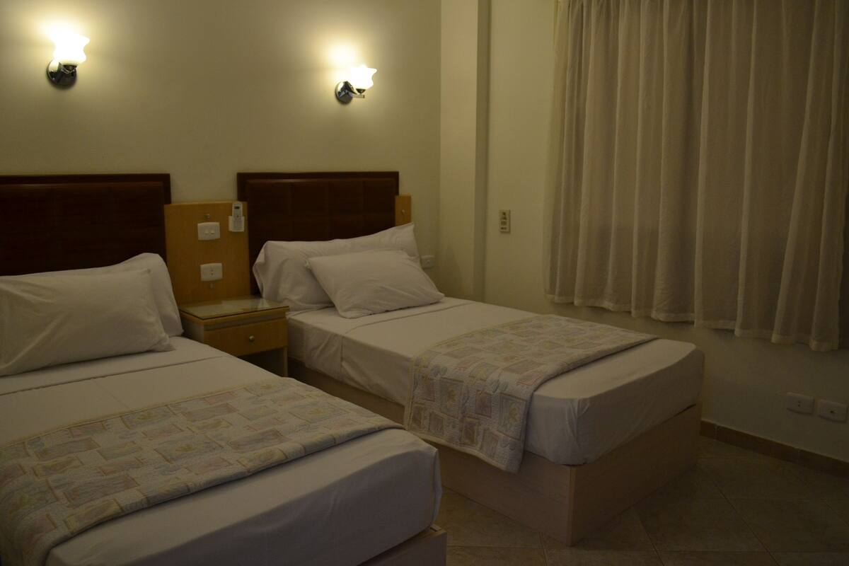 A hotel room with two single beds, a nightstand, a lamp, and a curtain.