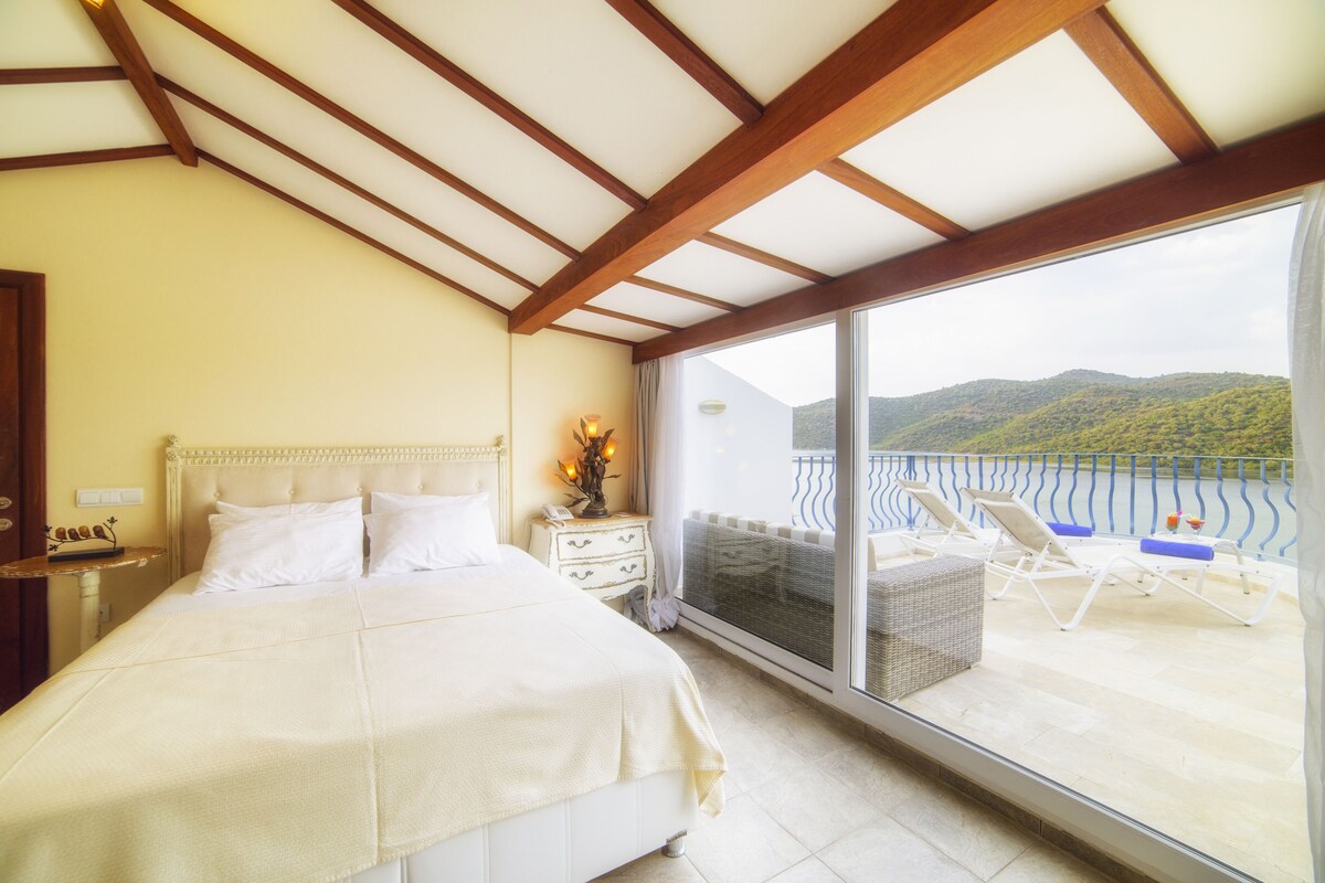 A bedroom with a large bed, a nightstand, a dresser, and a view of a balcony with lounge chairs.
