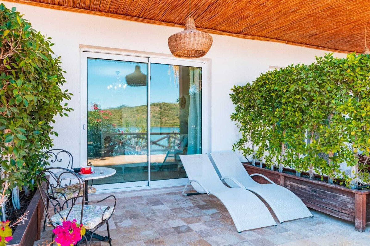 A sunlit patio with a glass door leading to a pool, surrounded by lush greenery and a wooden ceiling.