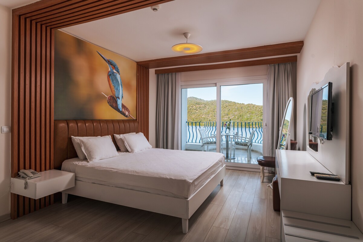 A modern hotel room with a large bed, a painting of a bird on the wall, a balcony with a view of the hills, and a TV on the dresser.