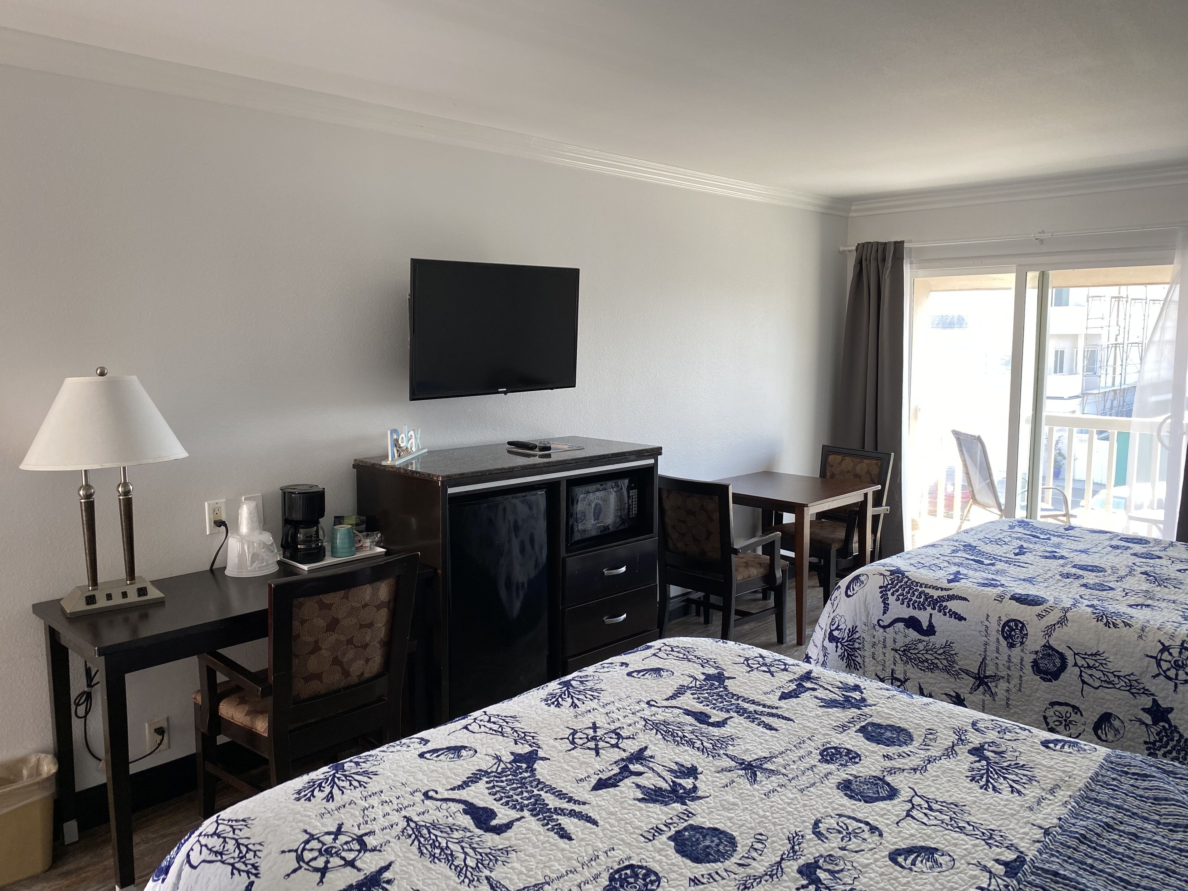 A hotel room with two beds, a desk with a lamp, a television, and a view of the outside balcony.