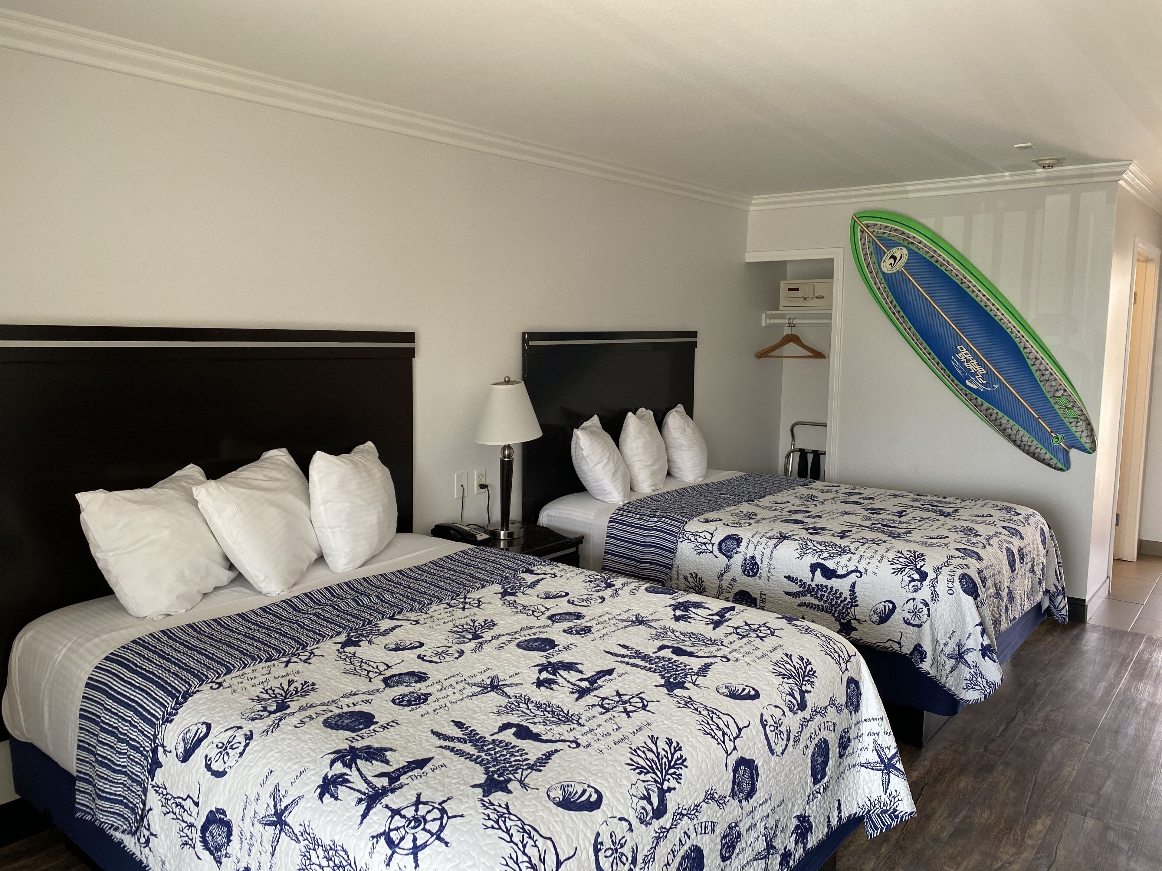 A hotel room with two beds, each with white bedding and blue floral patterns. There is a nightstand with a lamp and a phone on each bed. A surfboard is mounted on the wall.