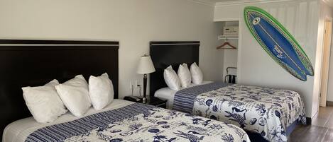 A hotel room with two beds, each with white bedding and blue floral patterns. There is a nightstand with a lamp and a phone on each bed. A surfboard is mounted on the wall.