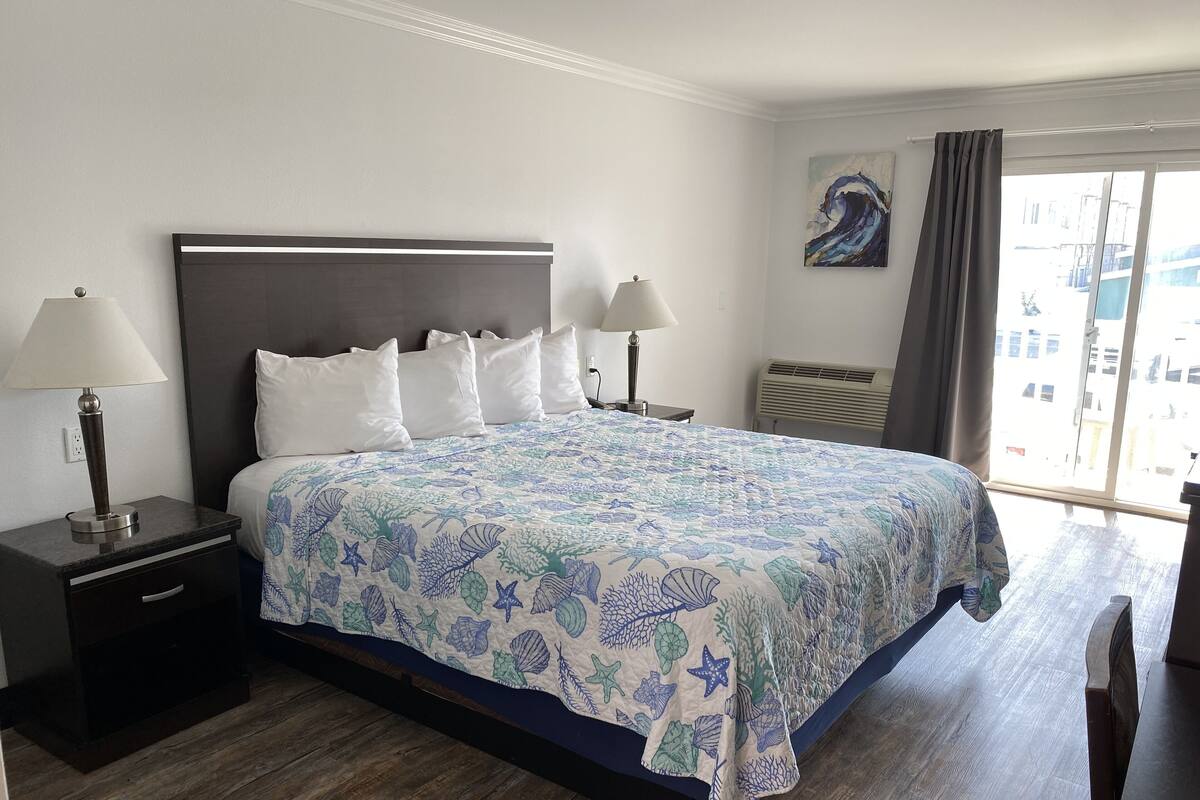 A hotel room with a large bed, two bedside tables with lamps, a nightstand, a window with curtains, and a painting on the wall.