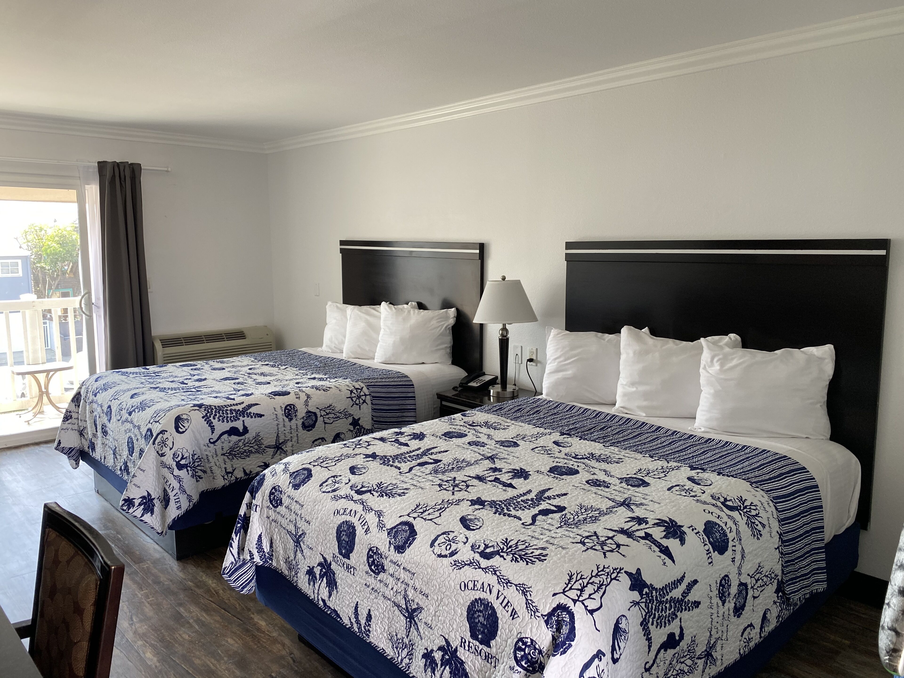 A hotel room with two beds, each with a dark headboard and white bedding. There is a window with curtains, a bedside table with a lamp, and a chair.