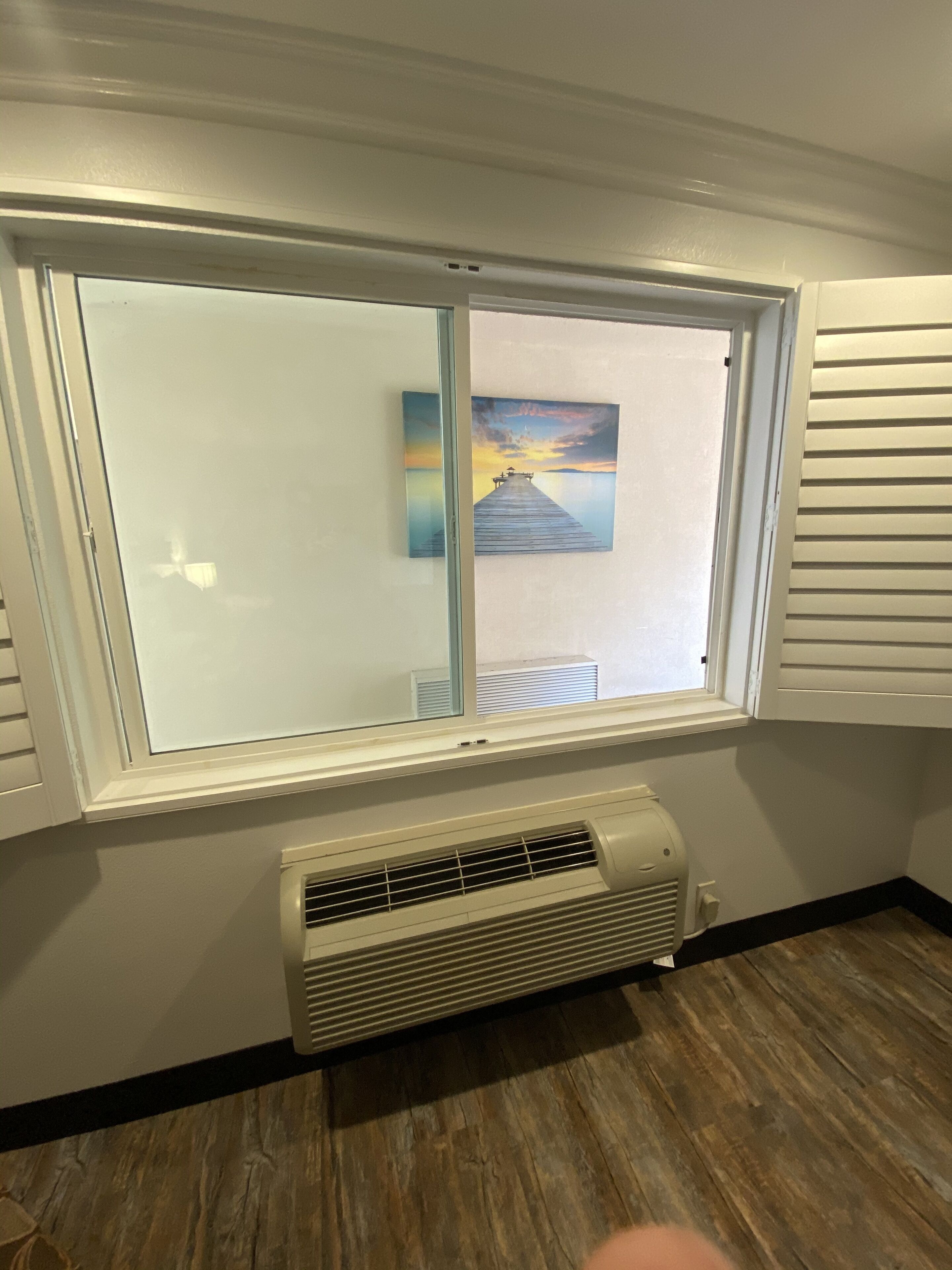 A room with a large window, a wall-mounted air conditioner, and a painting of a pier.