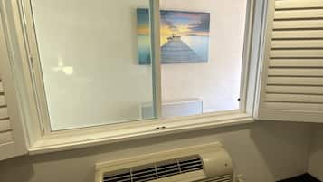 A room with a large window, a wall-mounted air conditioner, and a painting of a pier.