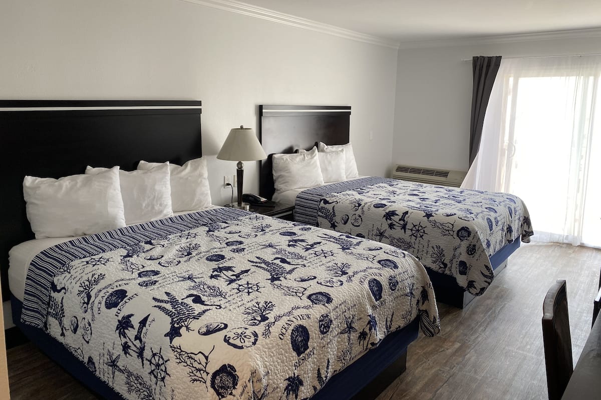 A hotel room with two beds, each with a headboard, white bedding, and a blue and white patterned bedspread. There is a bedside table with a lamp and a phone, and a window with curtains.