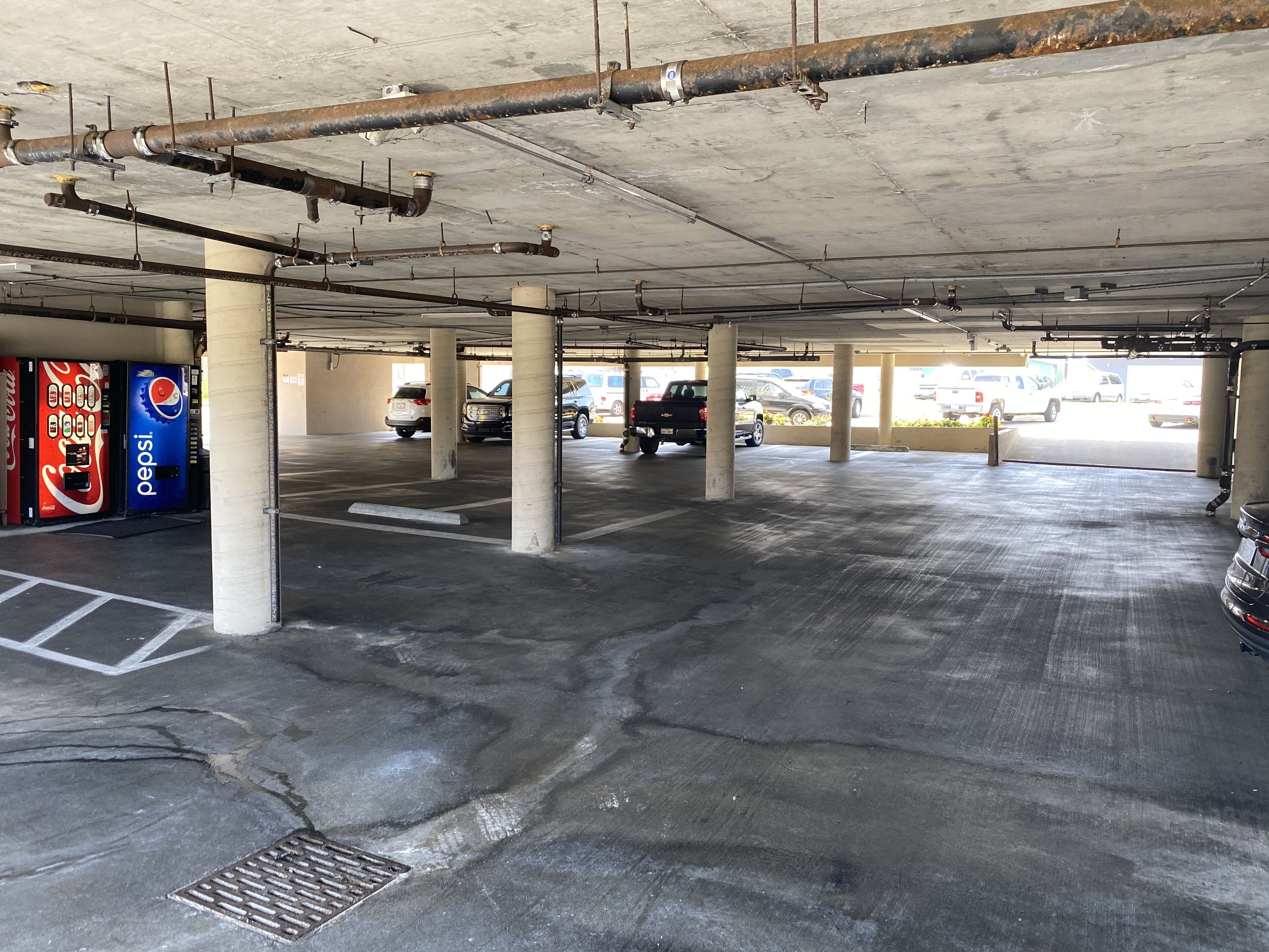 Un garaje para estacionamiento con pilares de hormigón, una máquina expendedora de Coca-Cola y Pepsi, y varios vehículos estacionados.