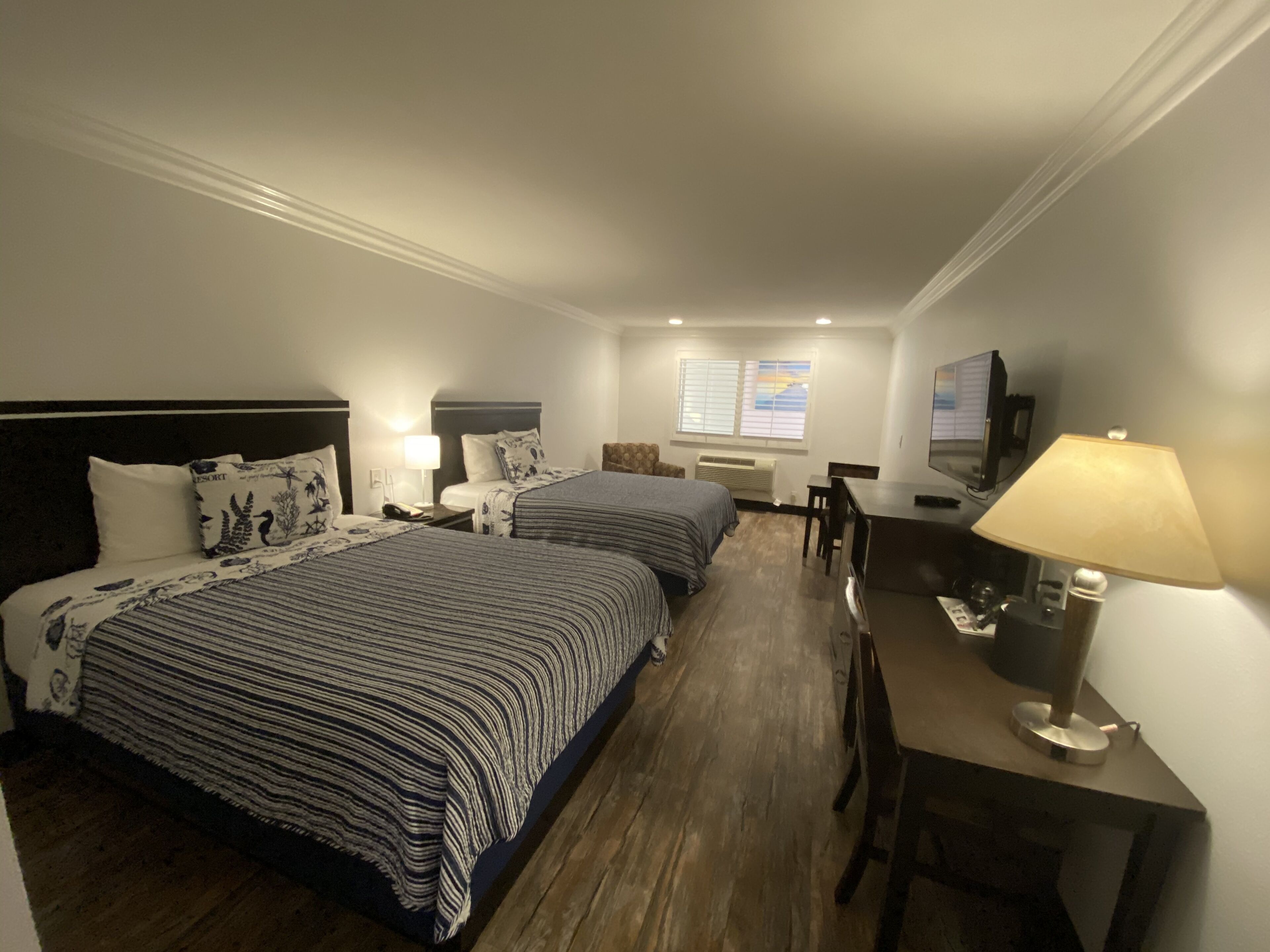 A hotel room with two beds, a desk with a lamp, a TV, and a window with blinds.