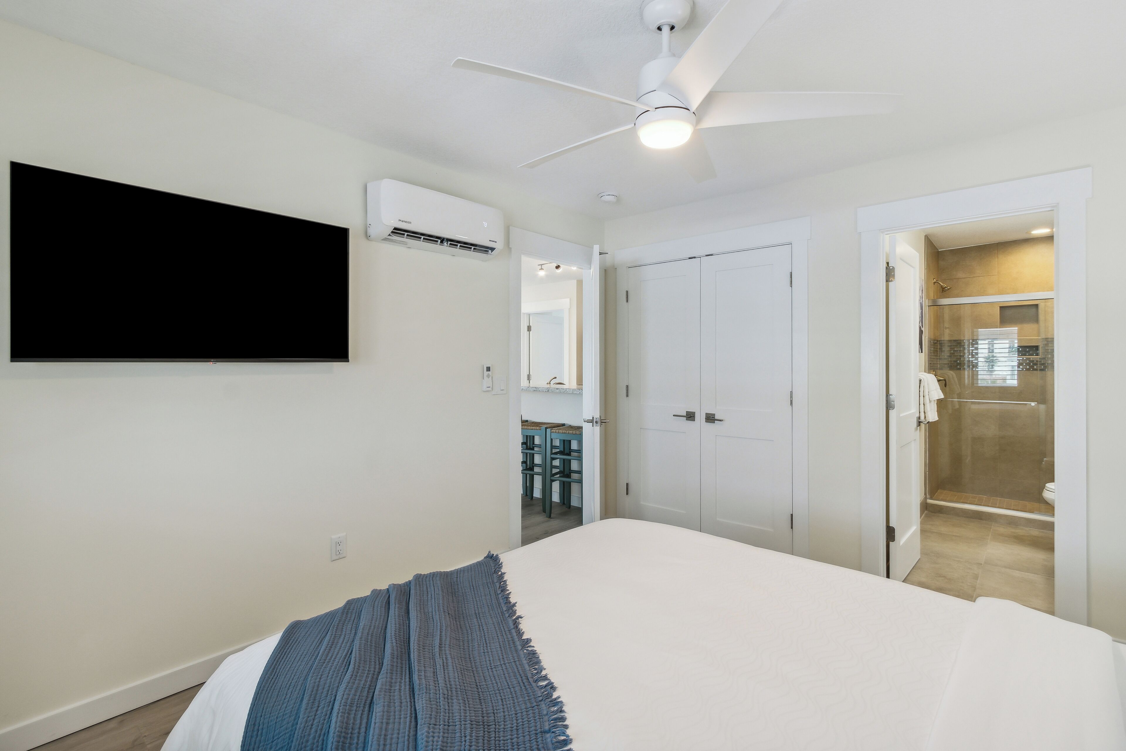 Un dormitorio con cama, ventilador de techo, equipo de aire acondicionado, televisor montado en la pared y un baño visible a través de una puerta abierta.