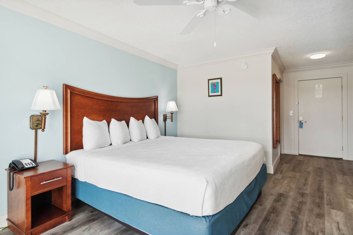 A hotel room with a large bed, wooden headboard, two bedside lamps, a nightstand with a phone, and a ceiling fan.