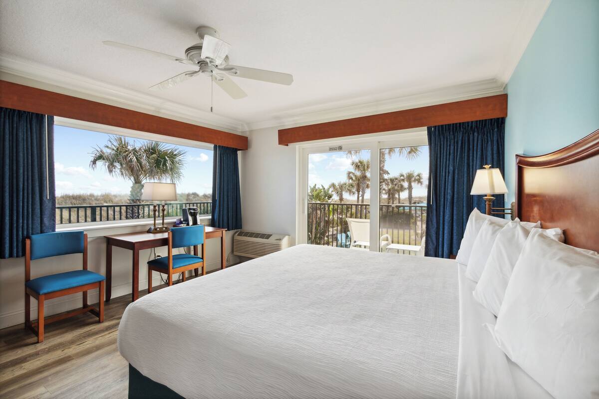 A hotel room with a large bed, a desk with a chair, a ceiling fan, and a view of palm trees through the balcony doors.