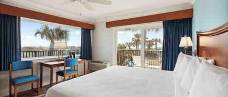 A hotel room with a large bed, a desk with a chair, a ceiling fan, and a view of palm trees through the balcony doors.