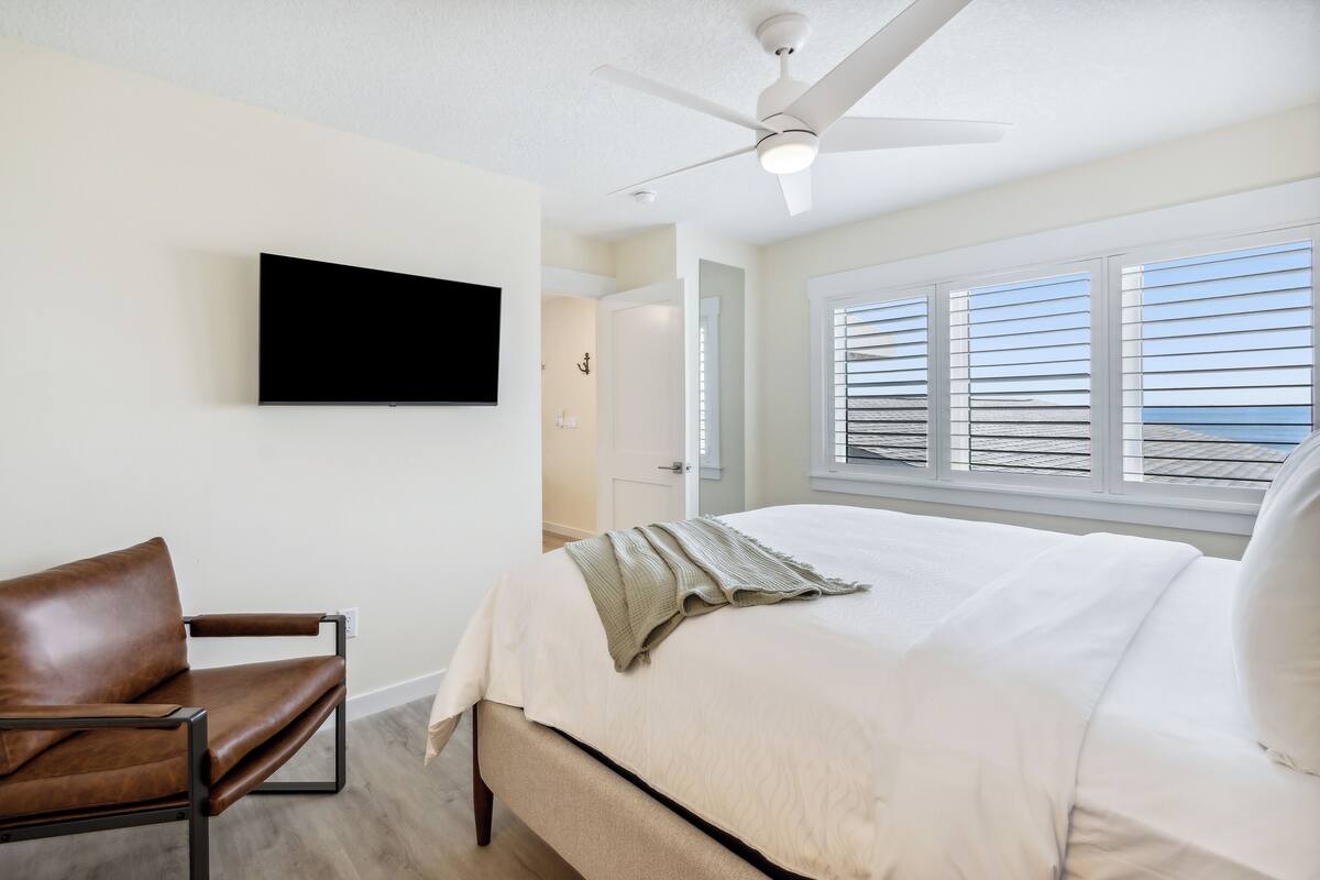 A bedroom with a bed, a brown leather chair, a ceiling fan, a TV mounted on the wall, and a window with blinds.