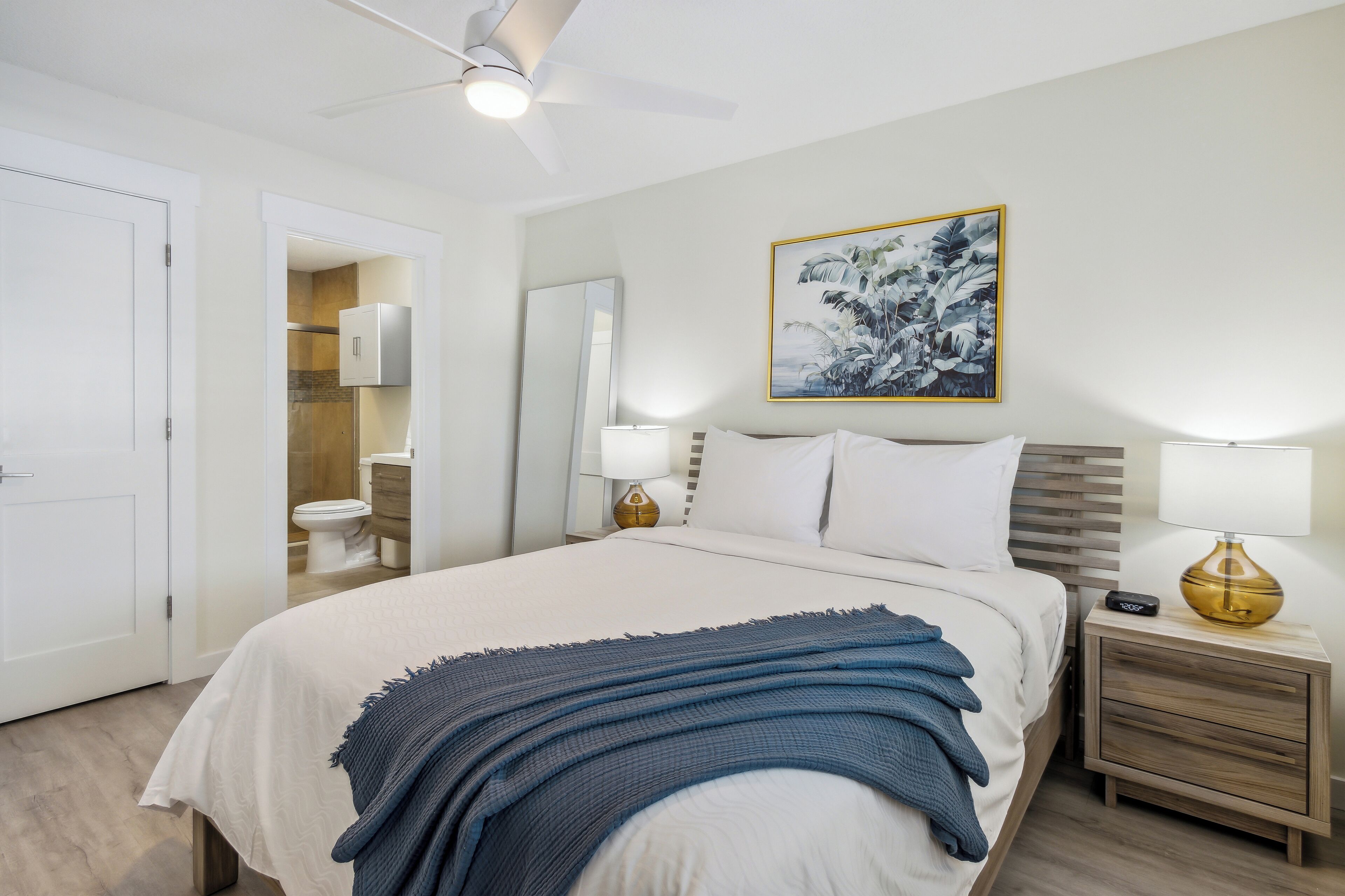 A hotel room with a bed, bedside tables, a ceiling fan, a bathroom with a toilet and sink, and a wall-mounted mirror.