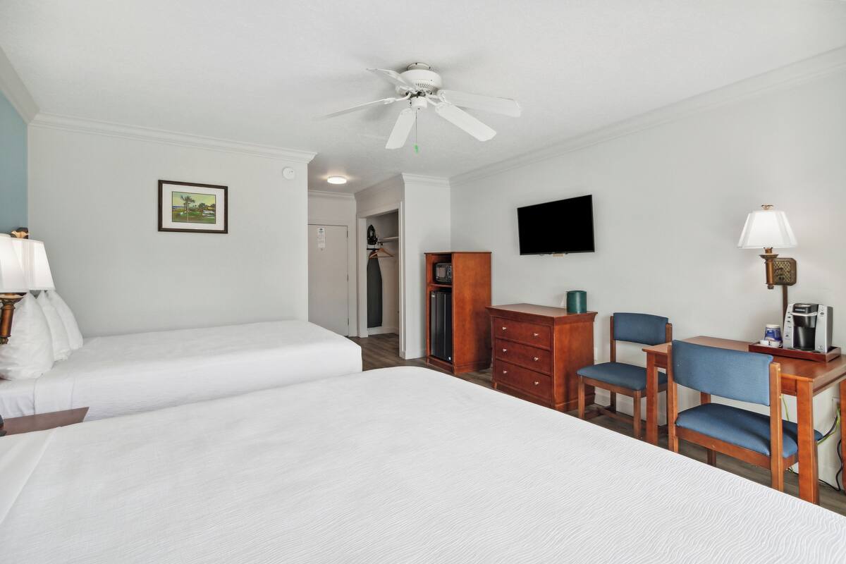 A hotel room with two single beds, a desk with a chair, a television, and a ceiling fan.