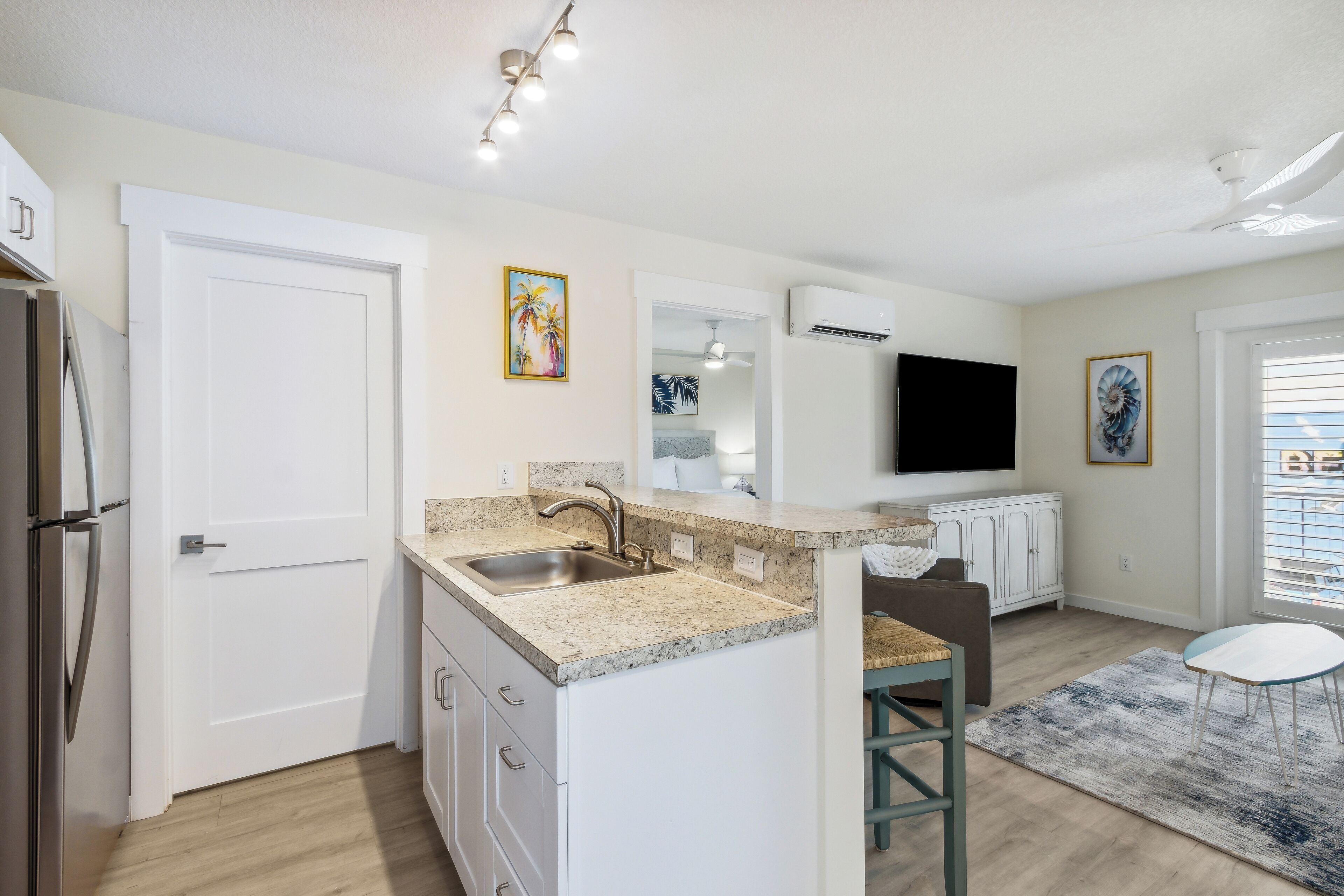 A modern kitchen with a granite countertop, stainless steel sink, and white cabinetry. There is a dining area with a table and chairs, and a large window with blinds.