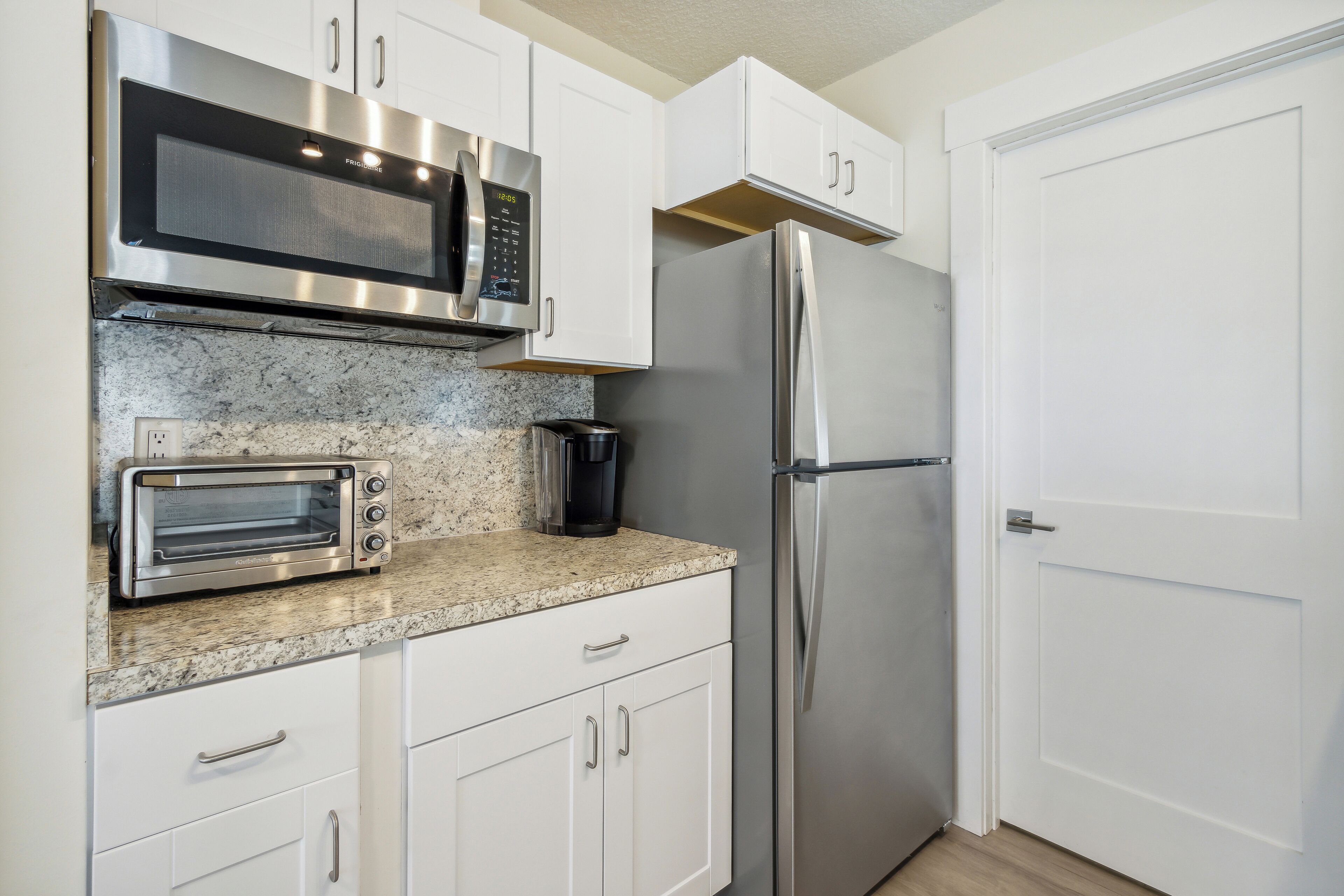 A kitchen with white cabinets, a stainless steel microwave, a toaster oven, and a refrigerator.