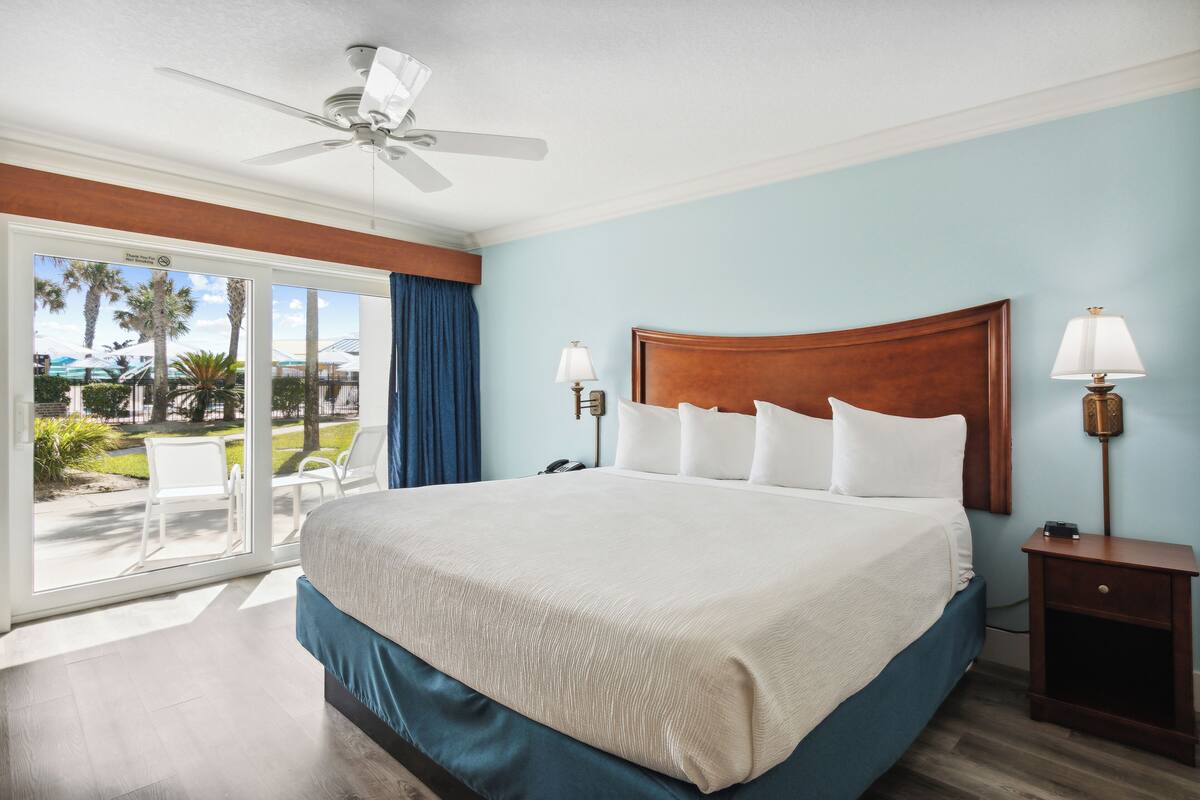 A hotel room with a large bed, a ceiling fan, a nightstand with a lamp, and a view of the outdoors through a sliding glass door.